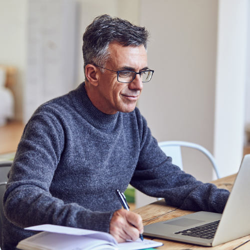 Hombre adulto, luce unas gafas con marco oscuro y buso gris, esta sentando frente al computador consultando y tomando nota de qué puede hacer y cómo puede planear su ahorro como complemento a su pensión obligatoria.