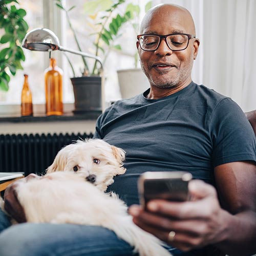 Sentado en su sofá, un hombre afrodescendiente calvo, con lentes y camiseta gris oscura, sostiene a su perro blanco mientras mira el celular