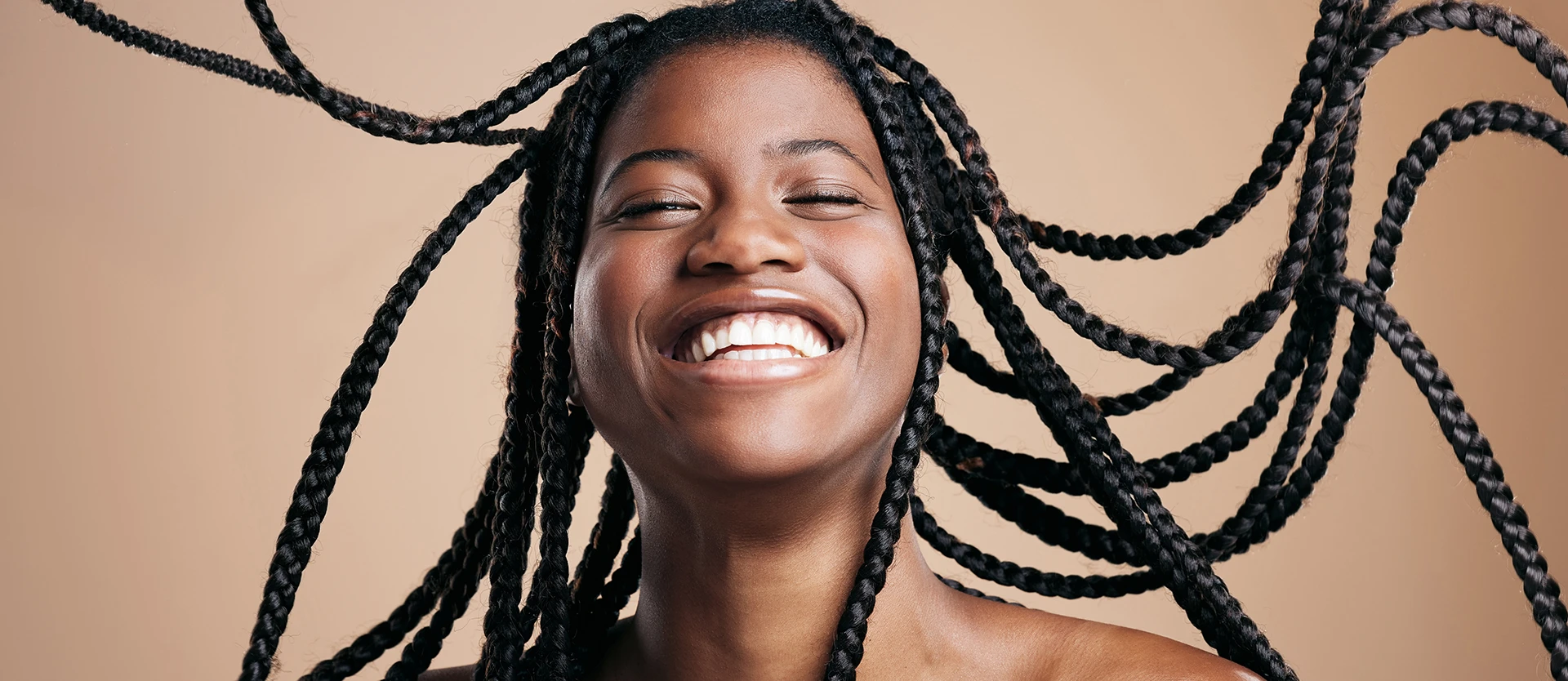 Smiling black woman with long flowing braids draped over her bare shoulders against a tan background.