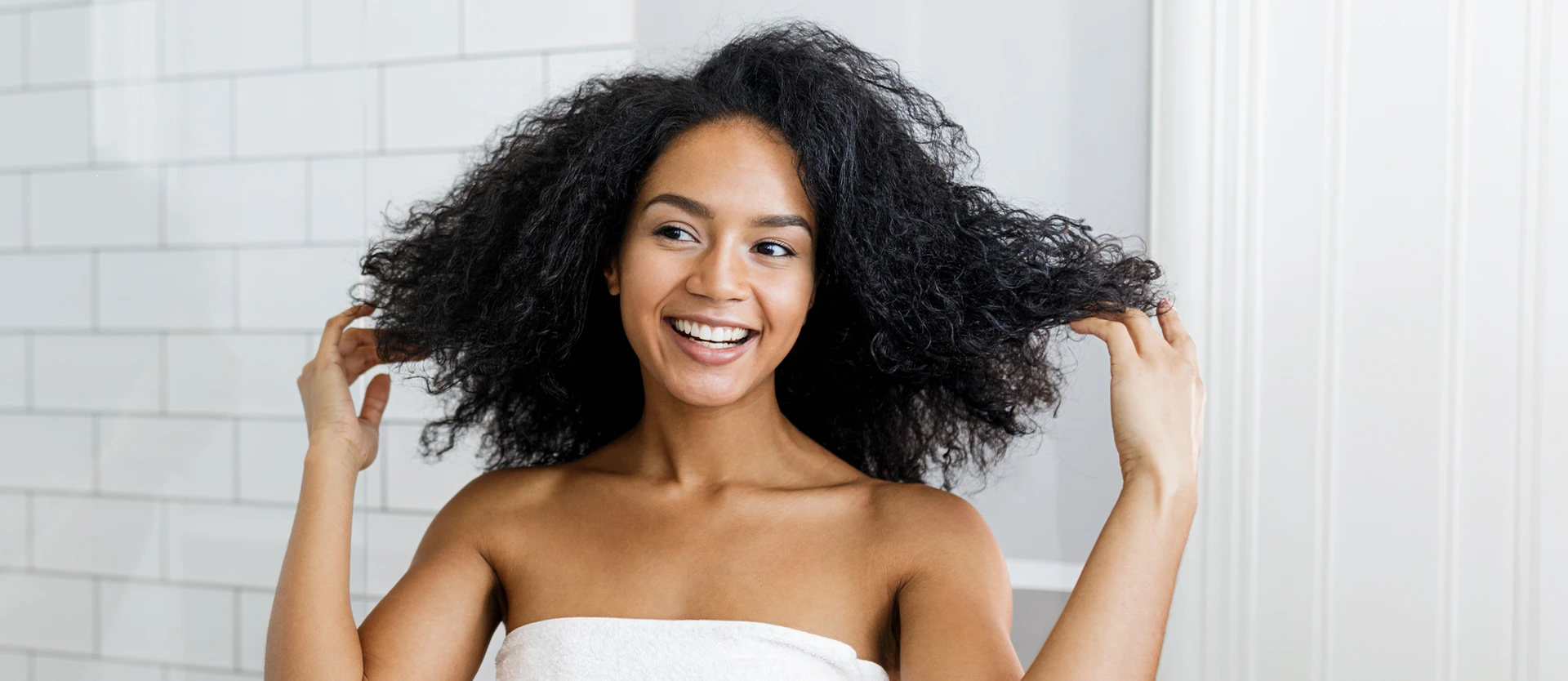 Young woman in a white towel with curly 4c type hair deciding on a hairstyle in front of a mirror