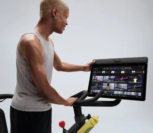 Man in a light gray tank top adjusts the touchscreen display on a Peloton Bike+