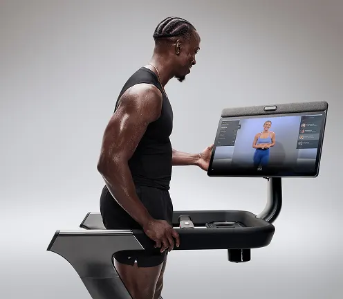 Man kneels on a mat in front of the Peloton Tread+ while adjusting the 23.8-inch swivel screen.