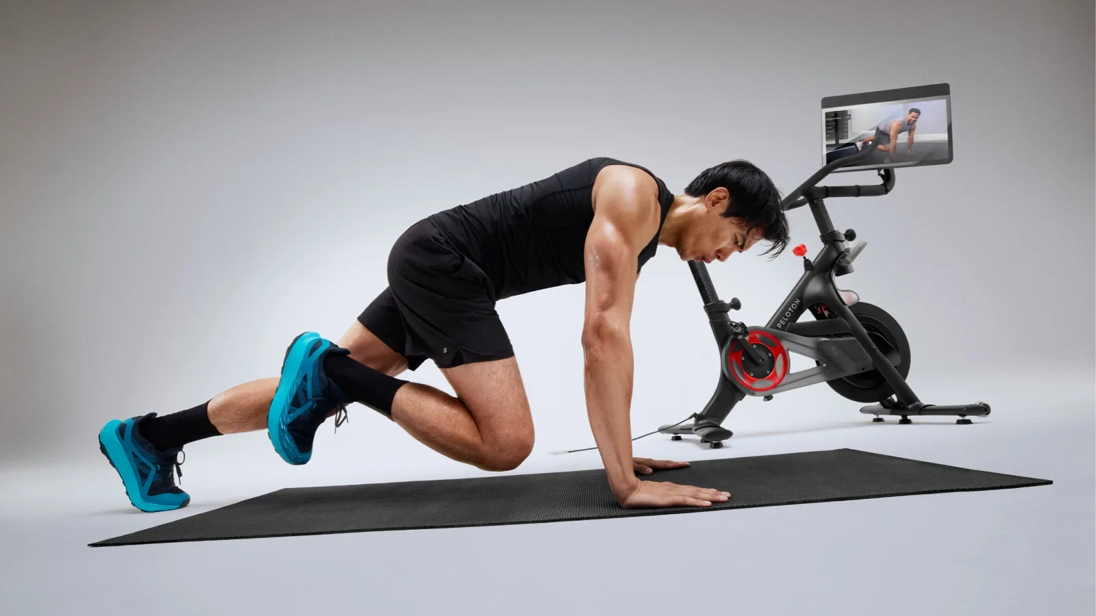 A man in dark clothing working out on a mat next to a Peloton Bike+ Pro