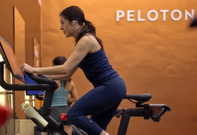 A woman in a Peloton Spaces environment on a Bike+ Pro. The Peloton logo is on the wall in the background.