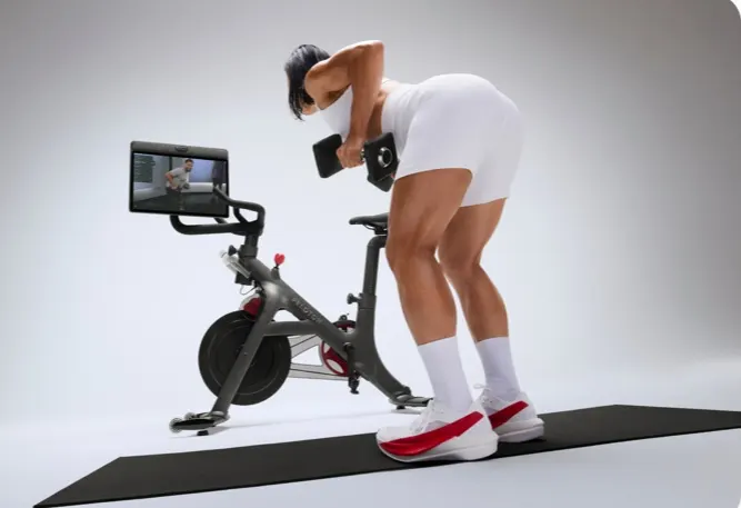 A woman working out with dumbbells on a mat next to Peloton Bike+ Pro. The Swivel Screen is turned so she can follow along.