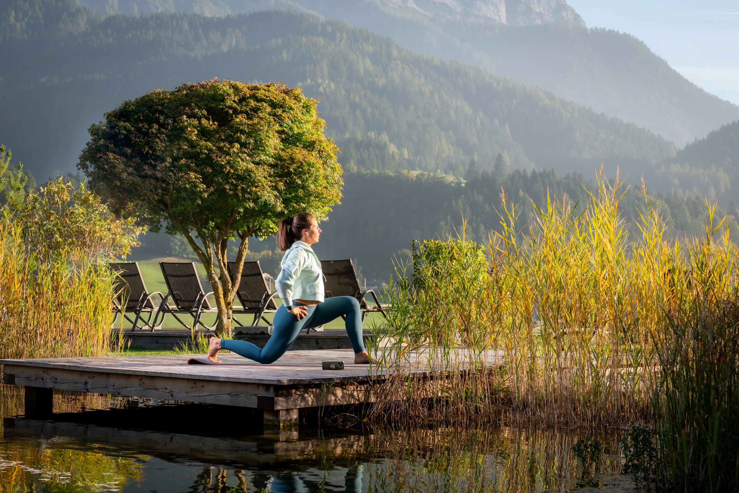 Hotelgast praktiziert morgens einen Peloton-Yoga Kurs am Teich auf dem Hotelgelände für einen entspannten Tagesbeginn.