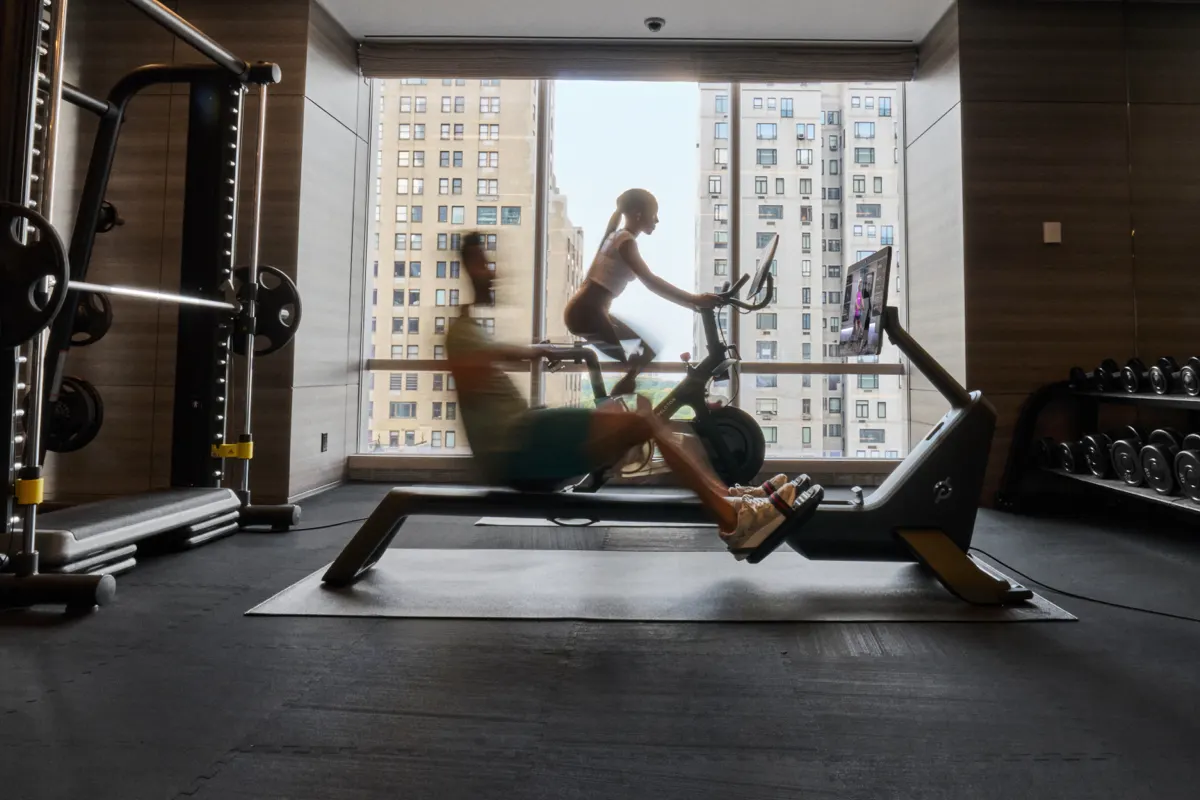 Two people working out in a gym with a cityscape outside of the window. One is on the Peloton Bike+ Pro and one is on the Peloton Row+ Pro.