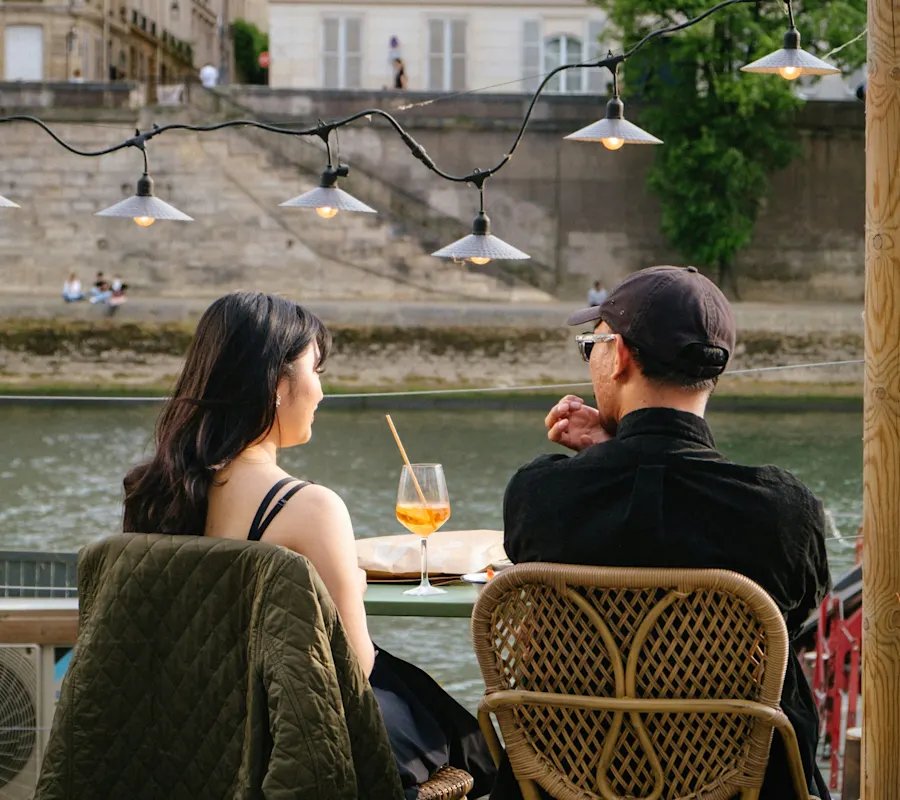 small couple, girl, woman and man on cafe chairs by water in city
