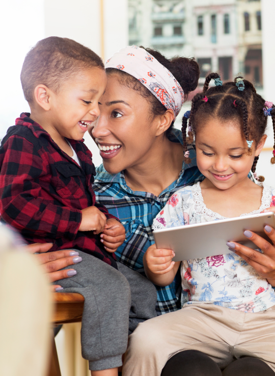 Femme qui tient ses deux enfants, une fille et un garçon, dans ses bras. La fille regarde l’écran d’une tablette.