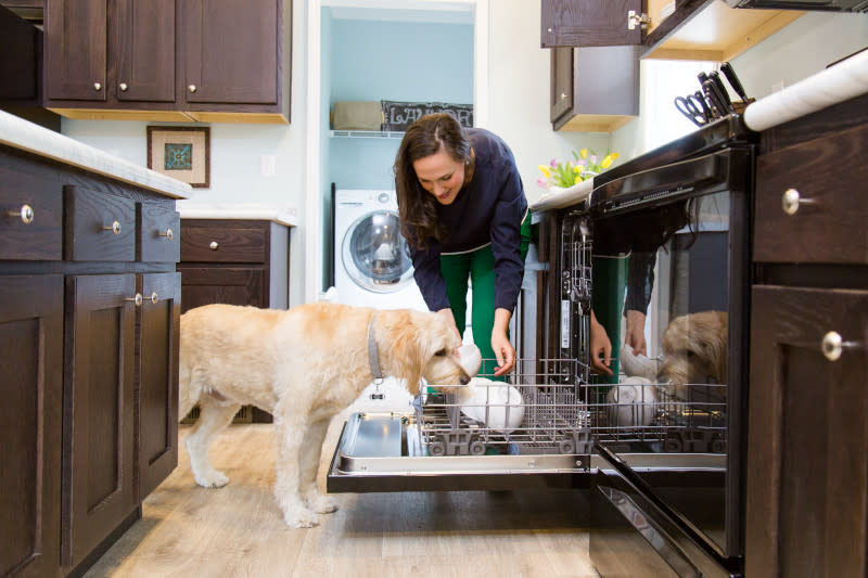 A smiling woman bends down to put a white bowl in the open dishwasher while her dog smells the bowls curiously.