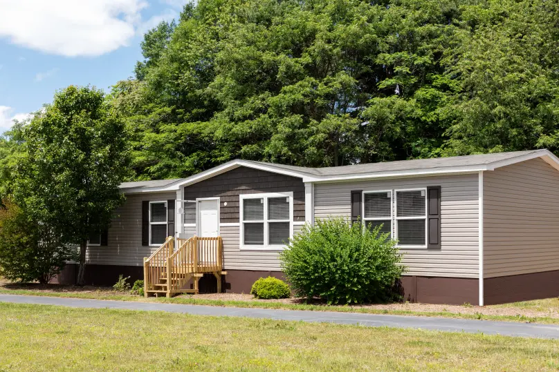 A brown and tan manufactured home is placed on a beautiful green plot of land on a sunny day. There’s a walkway in front of the home and a wooden staircase up to the home. 