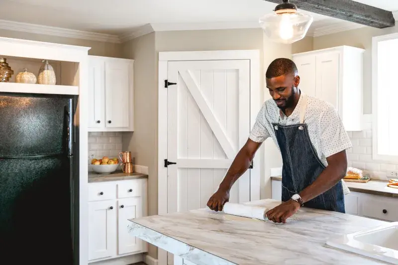 A man wears an apron and rolls dough on his kitchen island with white cabinets and a black fridge behind him.