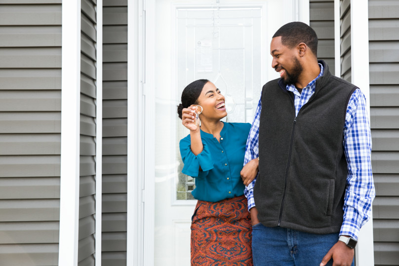 A young couple standing in front of a house laughing while the woman holds up house keys.