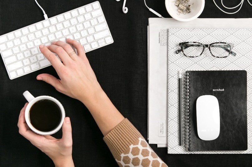A person's hands are seen holding a white coffee cup with the left and typing on a white keyboard with the right beside a pile of papers, glasses, a black notebook and a white wireless mouse.