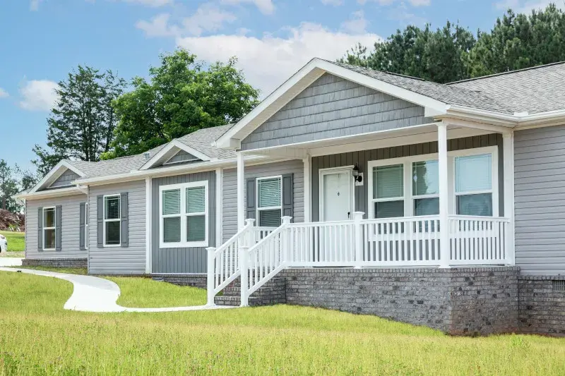 The exterior of a gray manufactured home with brick skirting, a front porch with white railing and a green lawn.