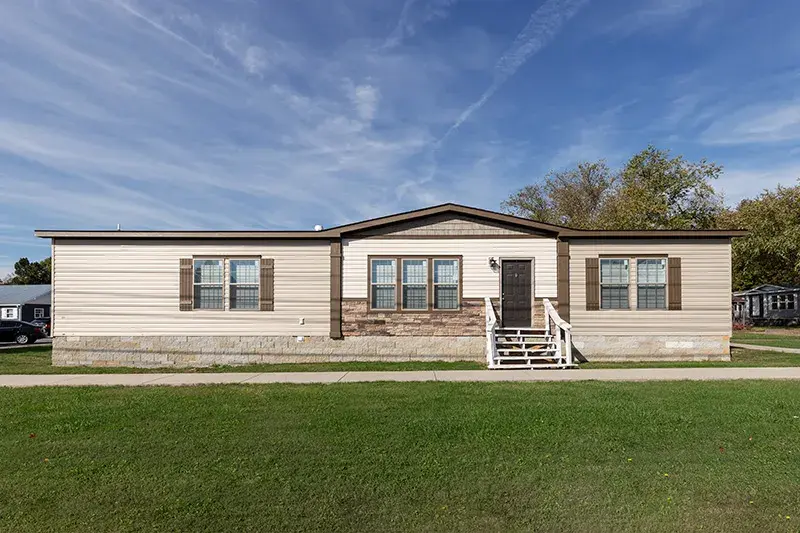 A manufactured home with beige and stone siding on green lawn and under a deep blue sky.