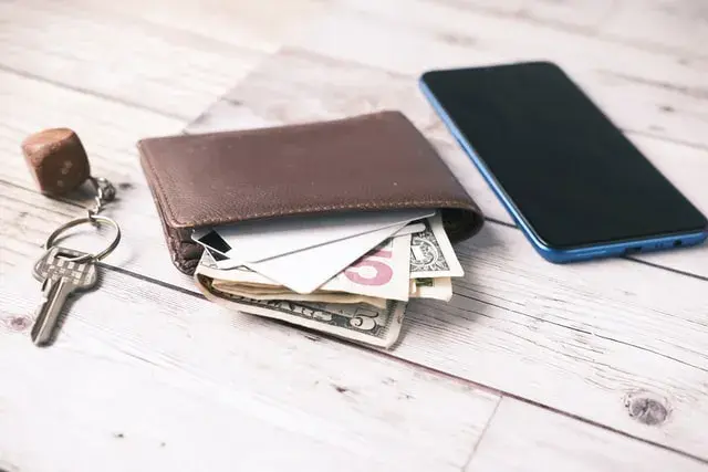 A leather wallet with dollar bills seen poking out of it is on a wooden surface with a cell phone on its right and a house key on a keychain on its left.