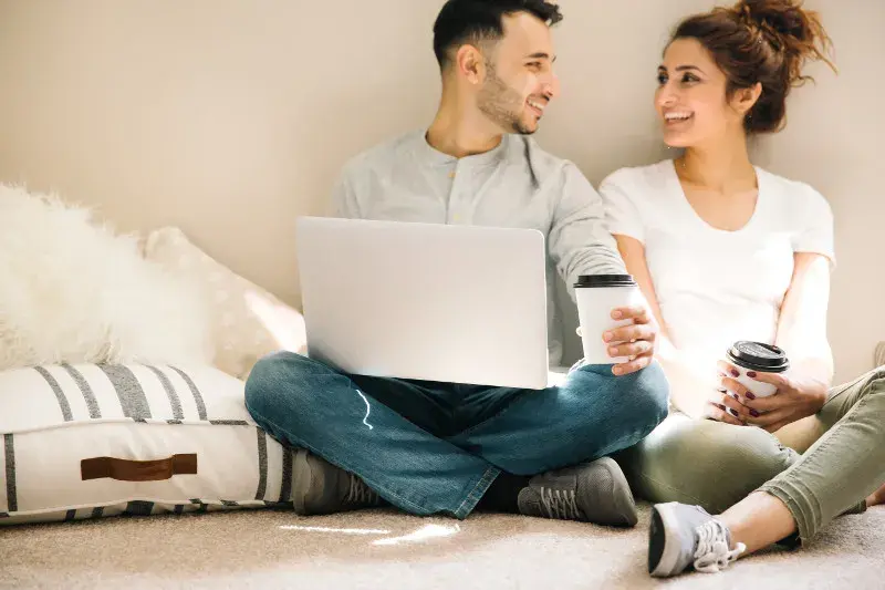 A happy couple smiles at each other after completing their online home loan application on the laptop in one of their laps.