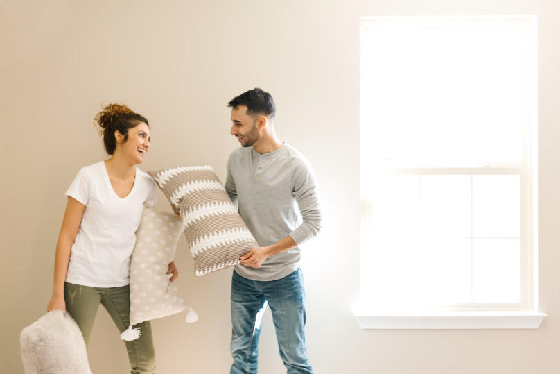 A young couple laughing and holding throw pillows in their new home.