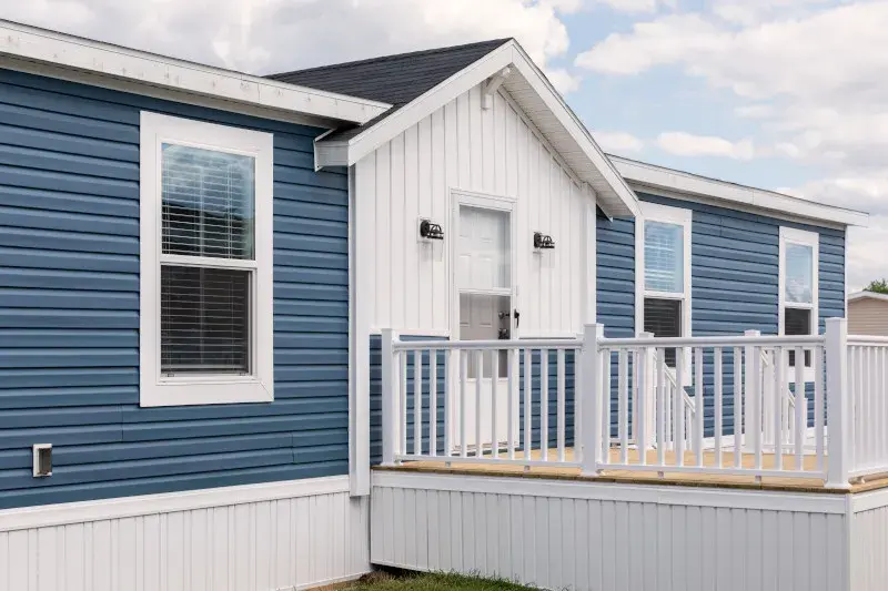 Exterior of home with a front deck attached. Home with white around the doorway and blue elsewhere on the home. It has a pitched roof with beam through the middle of the home. 