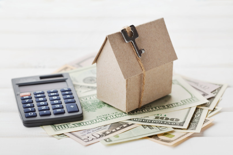 A calculator and a small house model are on top of scattered paper money.