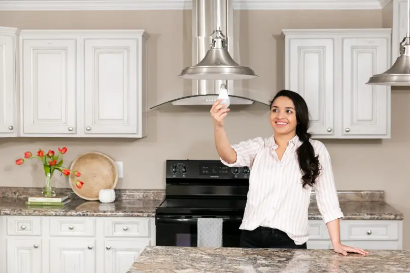 A woman in a striped shirt holds up an LED light bulb to screw into a metal pendant light in her kitchen.