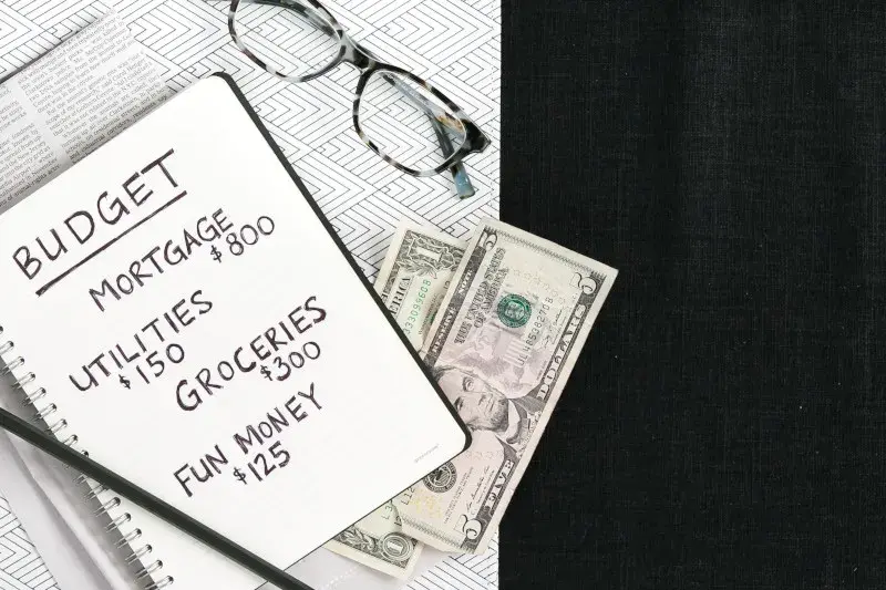 Photo shows a notebook with budgeting note, plus there is some loose cash and glasses also in view. 