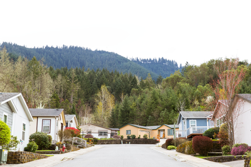 A shot of down the street of a neighborhood of manufactured homes with forested hills behind them.