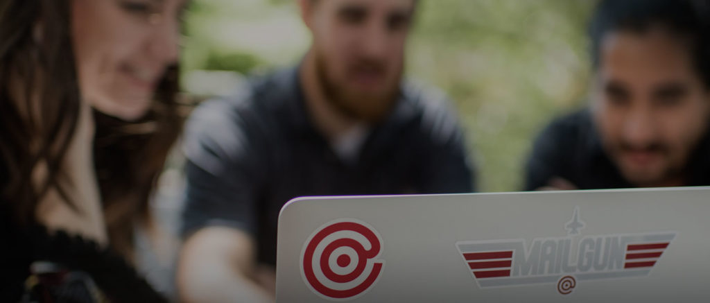 A photo of three people around a laptop with a Mailgun sticker on the back.