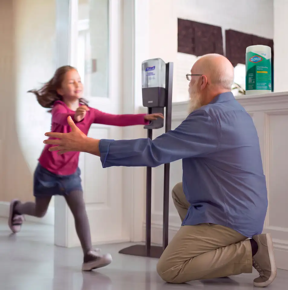 Young girl running to her grandfather in room for a hug, with Purell hand sanitizer dispenser and Clorox Disinfecting wipes on counter in background.