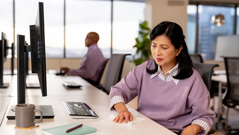 A woman in a lilac sweater wiping an office desk with a disinfecting wipe