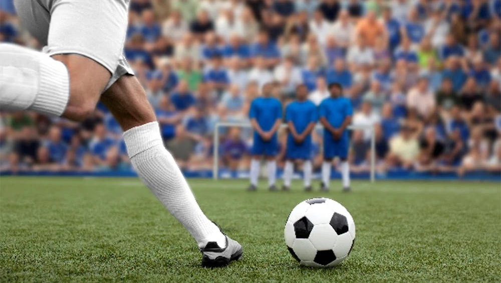 Legs kicking a soccer ball on a field towards 3 people standing together in front of a goal