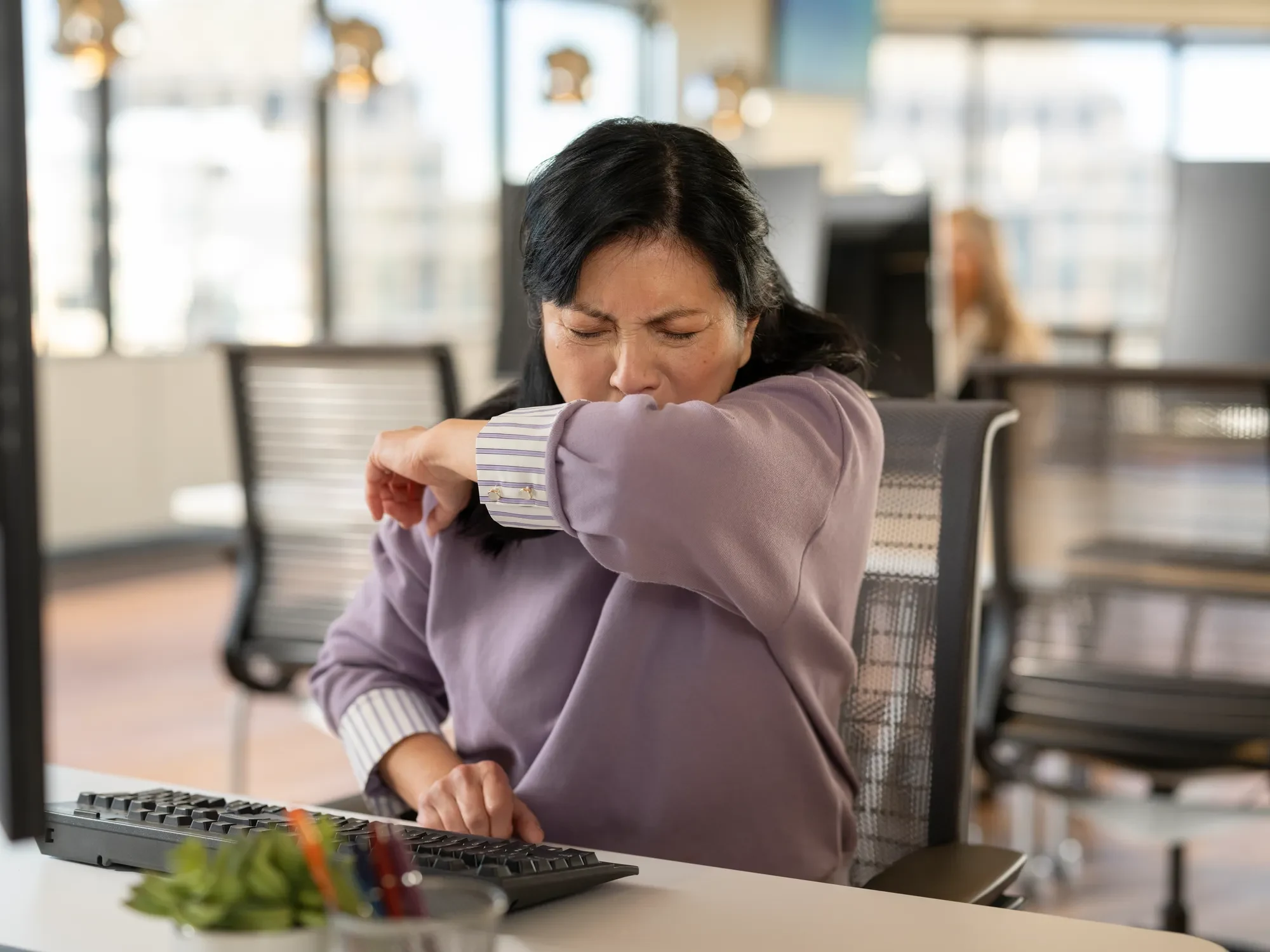 Woman sitting at desk in front of computer and covering her mouth with her arm as she sneezes.