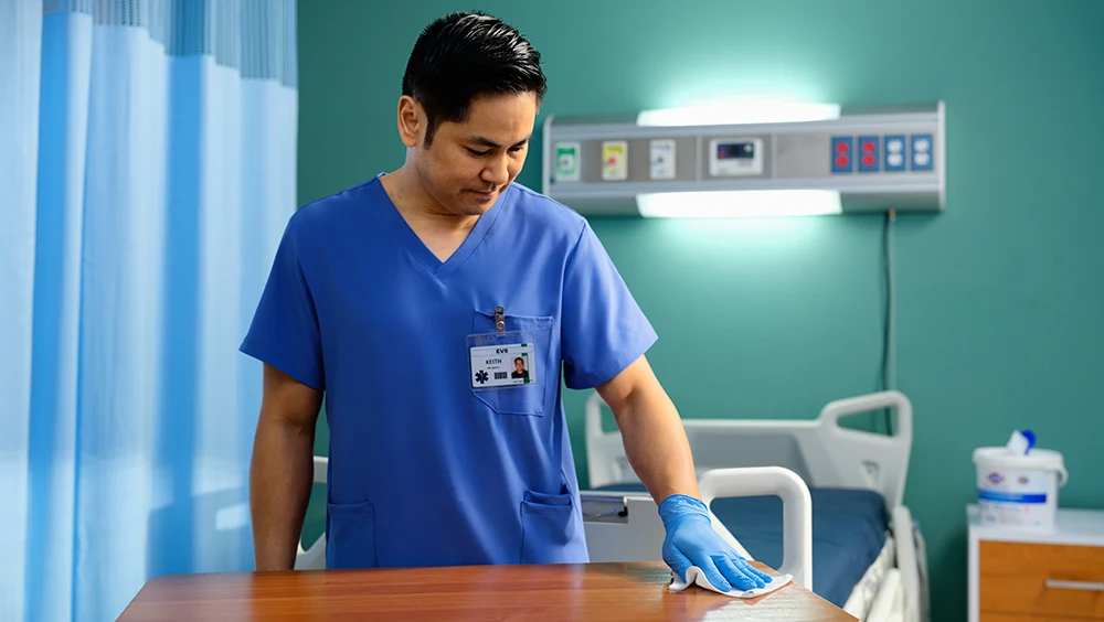 Man in scrubs cleaning a surface with wipes in a hospital room