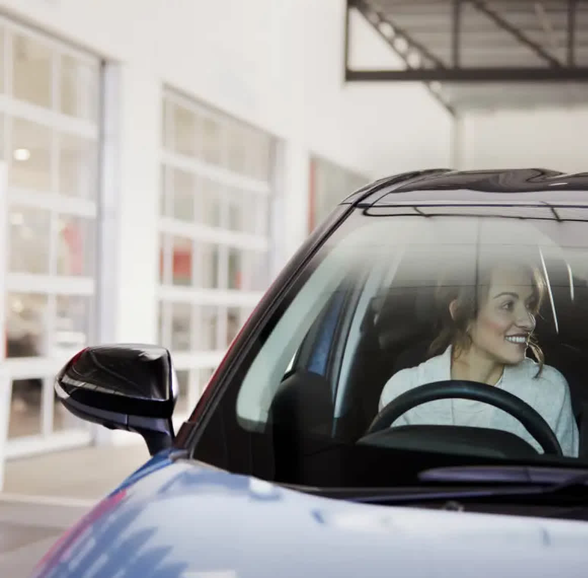 A woman sitting in a car on the driver's seat and smiling while looking to her left side.