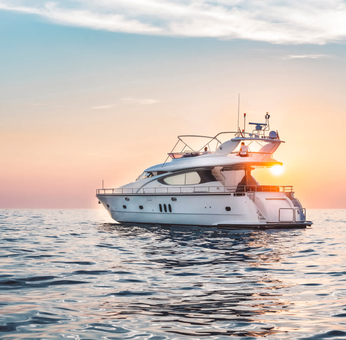 A small yacht floating in open water in front of pale blue and pink skies and the orange sunset appearing on the horizon in the background.