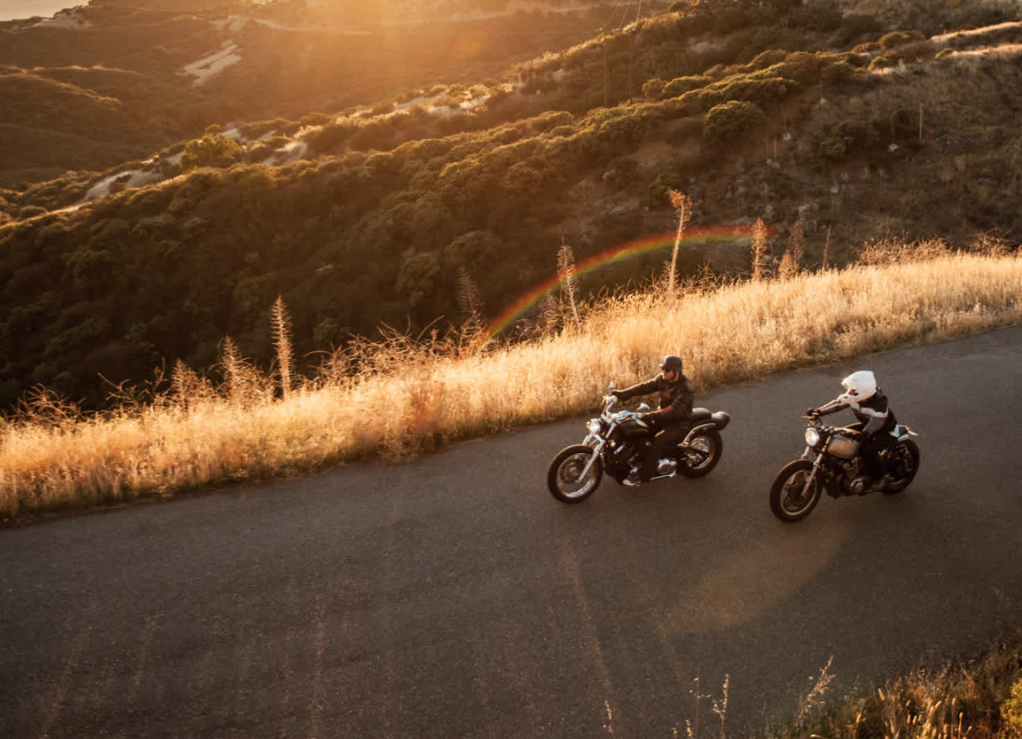 Two motorcycle riders driving down a road, overlooking a view of the late-afternoon sun over some mountains and fields.