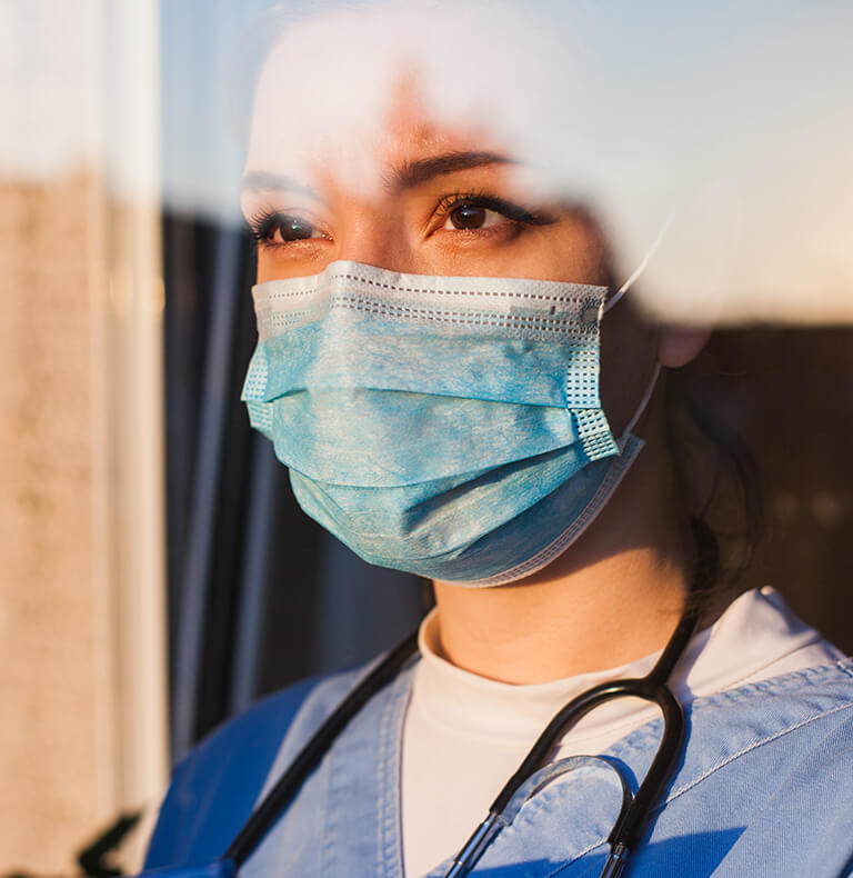 Frontline worker wearing a stethoscope and mask looks out at the sunset through a workplace window.