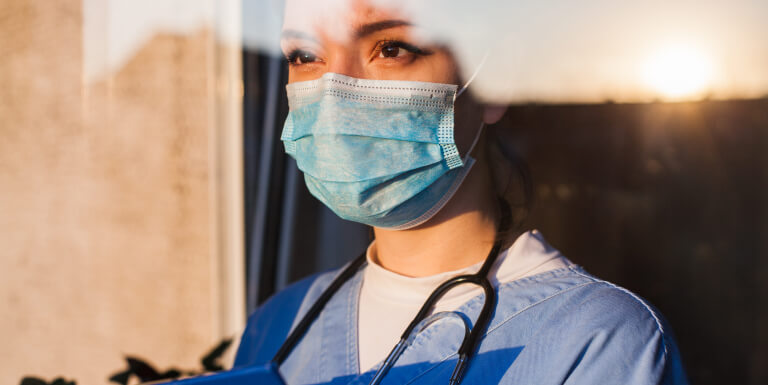 Frontline worker wearing a stethoscope and mask looks out at the sunset through a workplace window.