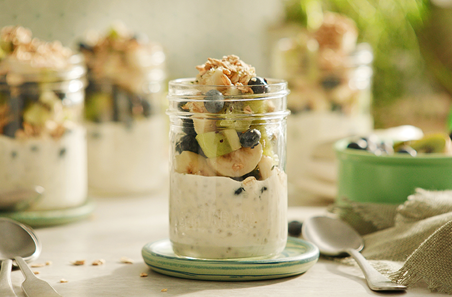 Mason jars filled with plant based yuzu citrus yogurt topped with sliced banana, kiwi, blueberries,  granola and chia seeds