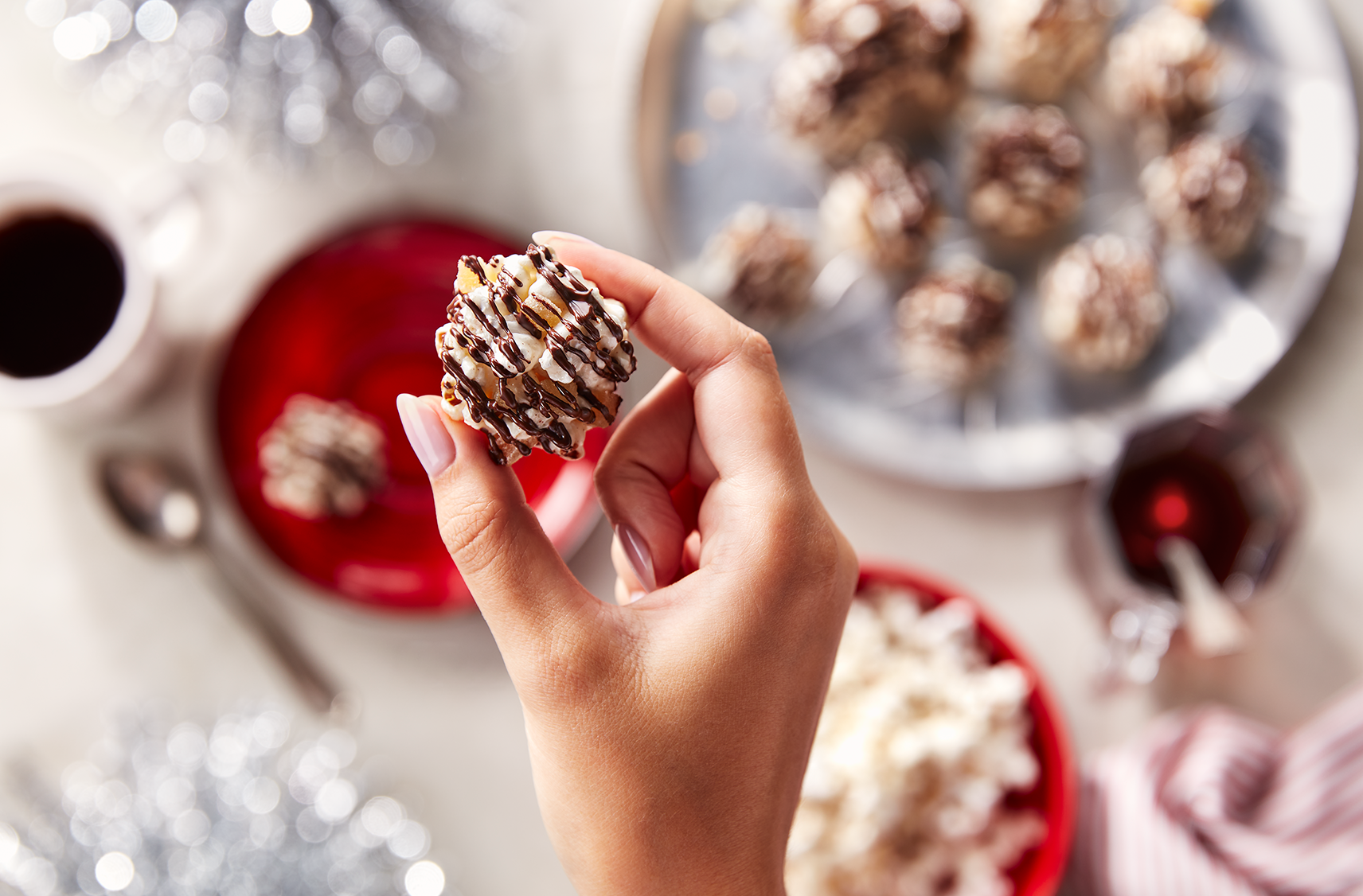 A dark chocolare gingerbread popcorn ball in a hand over a table top with the ingredients as well as a plate full ready made balls.