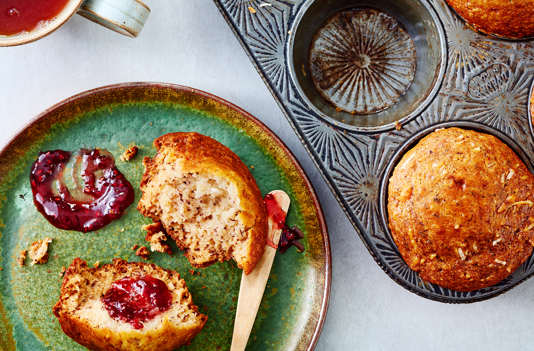 A split muffin spread with jam sits beside a pan of more muffins and a tea
