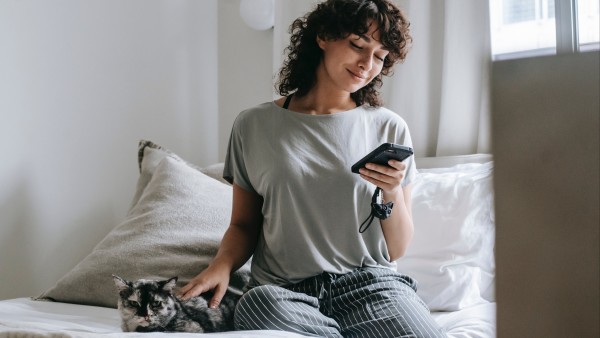 Smiling young woman using a smartphone and petting a cat on bed