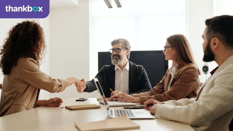 Man in black suit greets a woman in brown blazer with a handshake