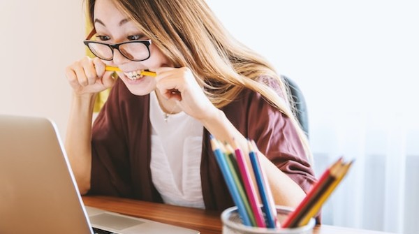 woman biting pencil while sitting on chair in front of computer during daytime