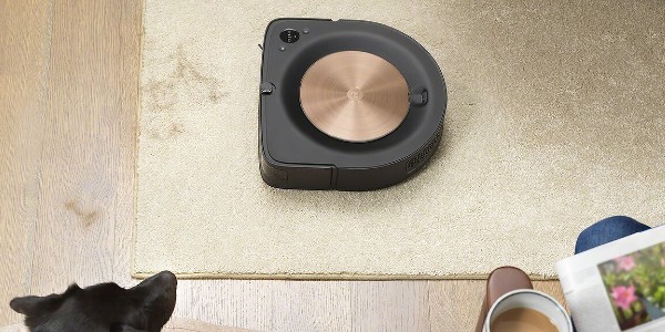 A Roomba vacuum on a rug with a dog and person enjoying coffee watching on