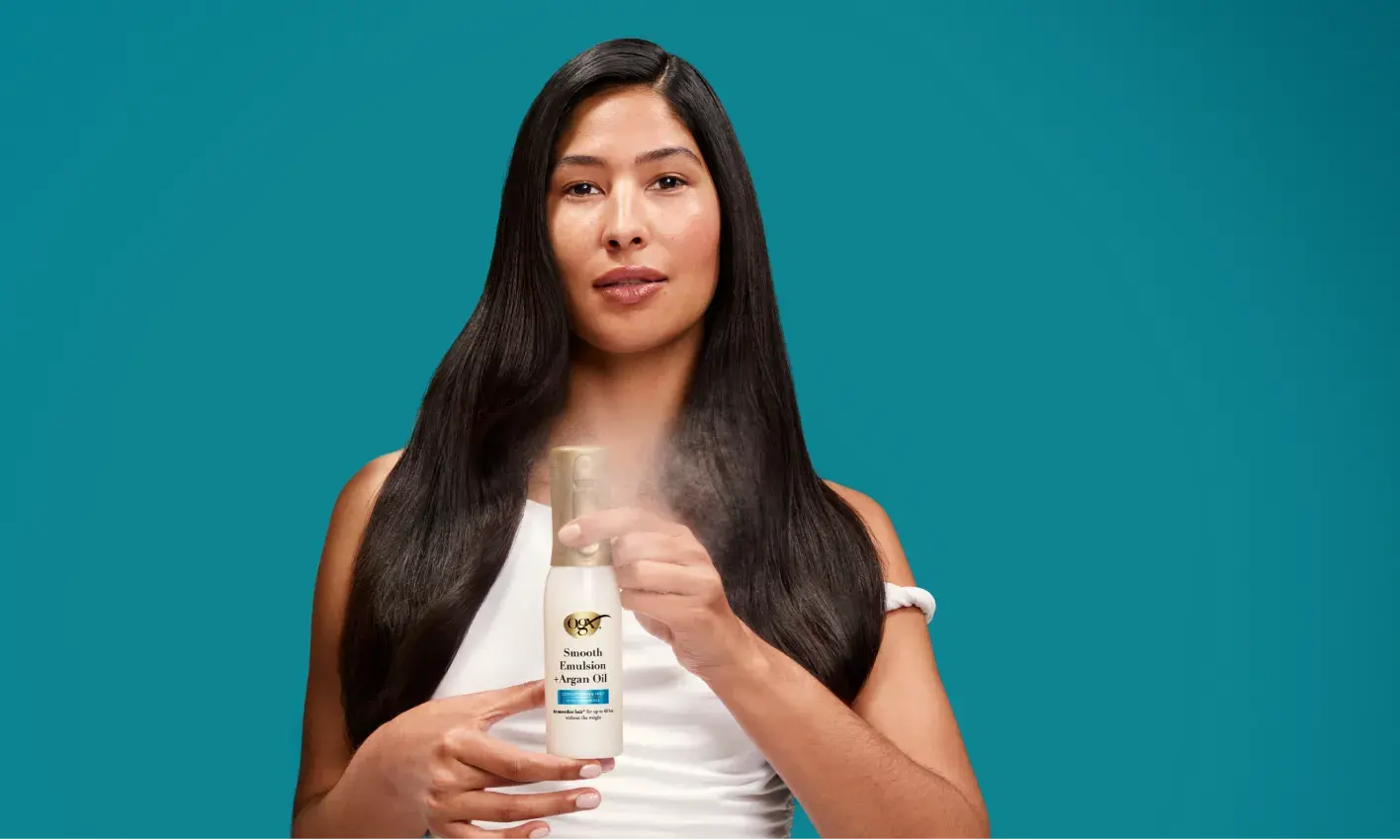 A woman with long, straight, dark hair is spraying a white bottle of "Smooth Essentials Argan Oil" hair product in front of her against a teal background.