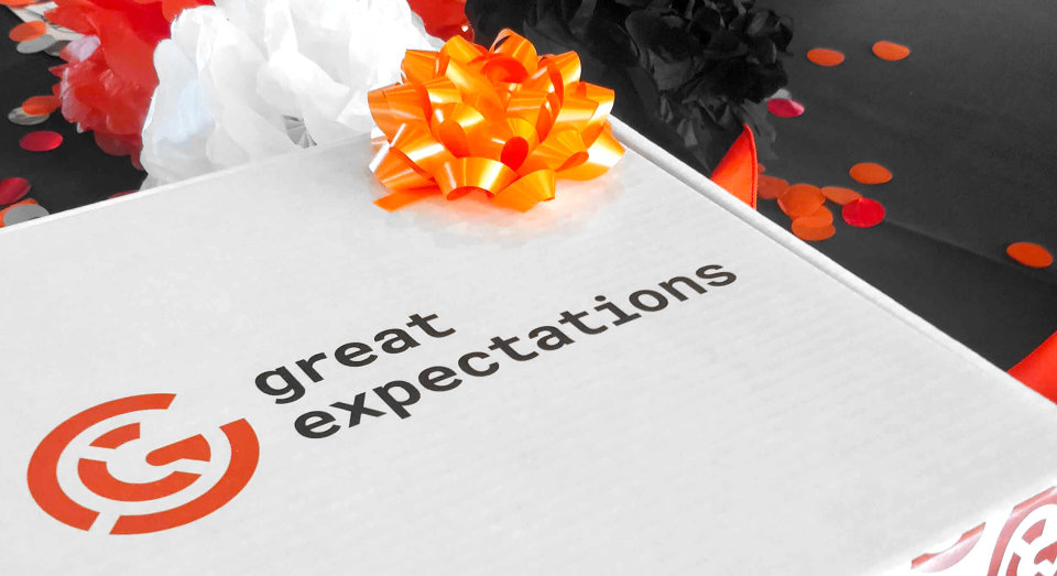 A white box with the new Great Expectations logo and an orange bow on top, with black, white, and orange decorations in the background