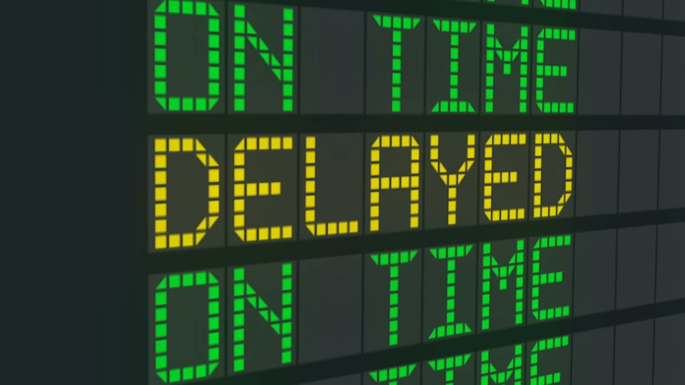 A photo of an airplane departure status screen centering a "delayed" status in a list of "on time" statuses