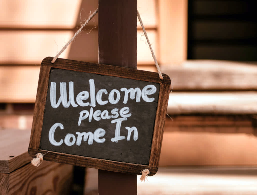 Photo of a chalkboard sign that says "Welcome please come in" on it hanging on a wooden post outside a building